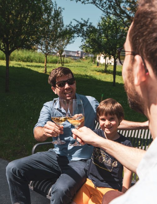 zwei Männer sitzen draussen in der Lounge auf der Terrasse bei sonnigem Wetter und stossen mit einem Weisswein an. Daneben sitzt noch ein Junge, welcher lächelt.
